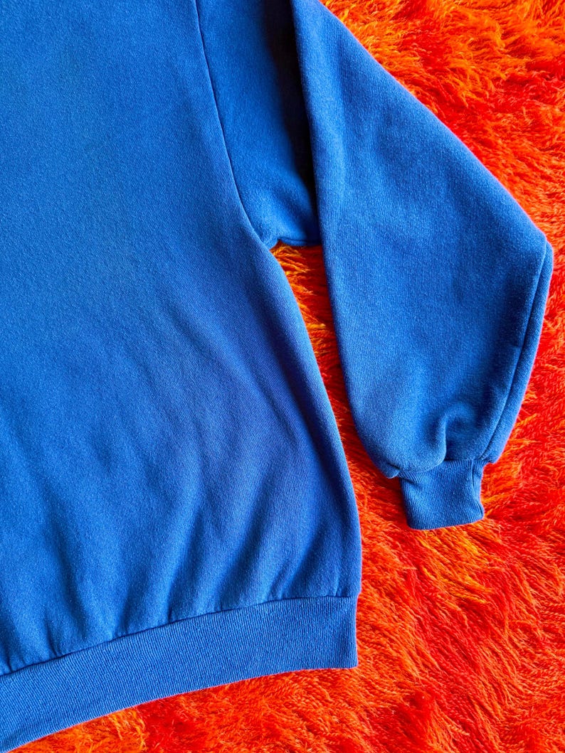 Pu&ograve; includere: Primo piano di una felpa blu reale. La felpa ha maniche lunghe con polsini e orlo a coste. Il tessuto sembra essere un materiale morbido e confortevole. Lo sfondo &egrave; una superficie arancione brillante e strutturata.