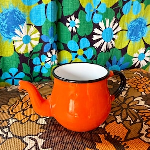 May include: An orange enamel teapot with a black handle and spout, set against a floral patterned background. The teapot has a white interior and a black rim. The background features a colorful floral design with blue, green, and white flowers.