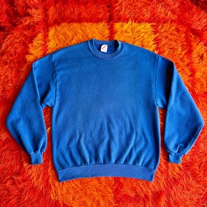 Pu&ograve; includere: Una felpa blu tinta unita con girocollo e maniche lunghe. La felpa ha polsini e orlo a coste. Lo sfondo &egrave; un vivace tappeto shag arancione e rosso. Il capo &egrave; realizzato in materiale morbido.