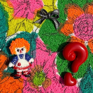 May include: A collection of vintage-style brooches and a figurine on a colorful floral patterned background. The items include a silver bow brooch, a red question mark-shaped pendant with a heart, and a doll figurine with orange hair and a blue dress.