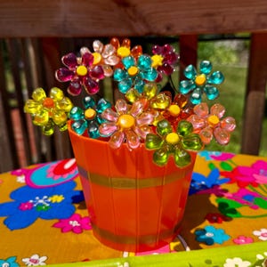 May include: A collection of translucent, multi-colored plastic flower-shaped decorations with yellow centers, arranged in a bright orange plastic bucket. The flowers feature petals in shades of pink, teal, green, and yellow, attached to thin stems.