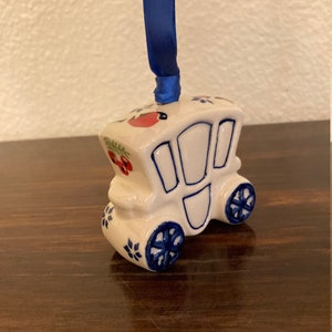 May include: A white ceramic ornament shaped like a horse-drawn carriage with blue and red painted details. The ornament has a blue ribbon attached to the top.
