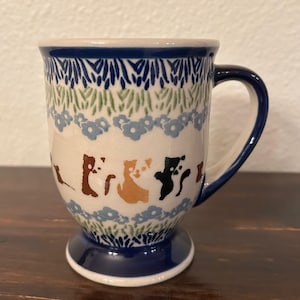May include: A ceramic coffee mug with a pedestal base. The mug features a blue rim and handle, with a cream-colored body decorated with blue, green, and brown floral and cat designs. The mug is approximately 4 inches tall.