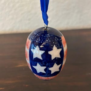 May include: A small, egg-shaped ceramic ornament featuring a glossy finish with a painted design of white stars on a dark blue field and red and white stripes. The ornament is suspended by a thin, bright blue ribbon attached to a metal loop.