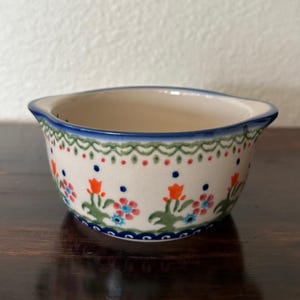 May include: A small, off-white ceramic bowl with a floral pattern. The bowl features a blue rim and a decorative border with red, green, and blue accents. The floral design includes orange, blue, and pink flowers.