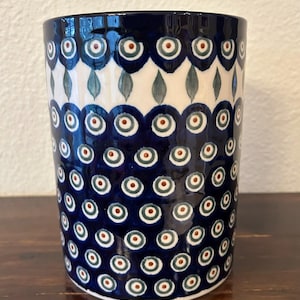 May include: A cylindrical ceramic mug featuring a dark blue glaze decorated with a repeating pattern of white and teal peacock eye motifs, accented with small brown centers, arranged in horizontal rows across the exterior surface.