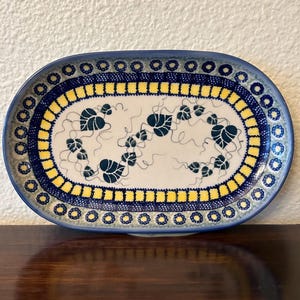 May include: An oval ceramic serving platter with a blue rim and a yellow and blue patterned border. The center features a white background with a leaf design in dark teal. The platter is on a dark wooden surface.