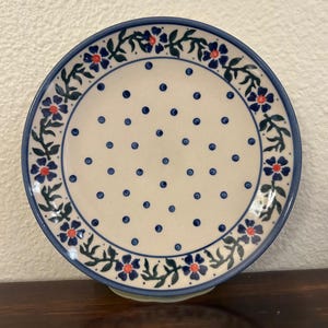 May include: A round ceramic plate with a cream-colored center and a blue rim. The plate features a pattern of blue dots and a border of blue flowers with red centers and green leaves. The plate is resting on a light green base.