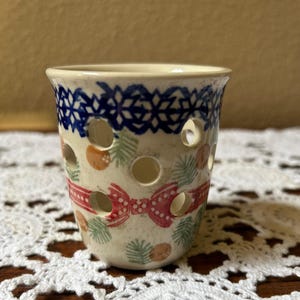May include: A ceramic candle holder with a cream base, adorned with a blue geometric pattern at the top and a red bow design. The holder features circular cutouts and is decorated with green and orange accents.