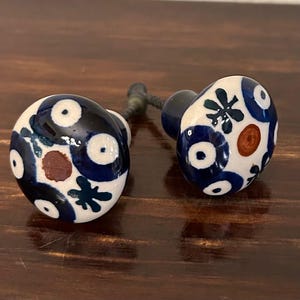May include: Two ceramic cabinet knobs with a white base, adorned with a blue floral pattern and brown accents. The knobs are attached to dark metal stems, set against a dark wood surface.