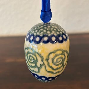 May include: A ceramic egg ornament featuring a hand-painted pattern of green swirling floral motifs on a yellow background, accented by blue circular bands at the top and bottom. The egg is suspended by a vibrant blue satin ribbon for hanging.