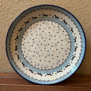 May include: A round ceramic plate with a blue rim and a white interior. The plate features a repeating pattern of black dachshund dogs and paw prints in blue and black. The plate has a decorative blue border.