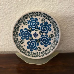 May include: A small, round ceramic plate with a white base and a blue floral design. The plate features five stylized blue roses and a border of blue and green leaves. The plate is approximately 4 inches in diameter.