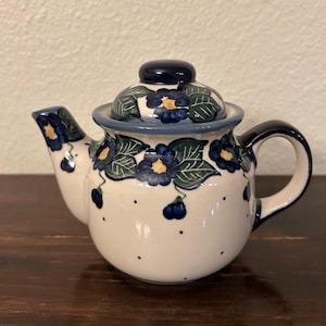 May include: A ceramic teapot with a white base, adorned with blue floral patterns and a matching lid. The teapot features a curved spout and handle, with a dark blue accent on the lid and handle. The floral design includes blue flowers with yellow centers and green leaves.