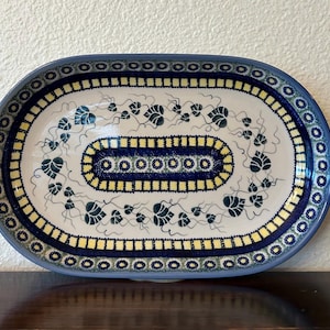 May include: Oval ceramic serving platter with a blue rim and intricate floral patterns. The platter features a white base with blue and yellow accents, and a central design of stylized leaves and vines. The overall design is symmetrical and decorative.