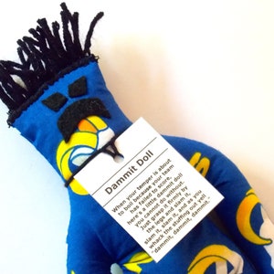 May include: A blue Dammit Doll with black yarn hair and a black felt mustache and eyebrows. The doll has a tag that reads "Dammit Doll" and instructions on how to use it when your temper flares.