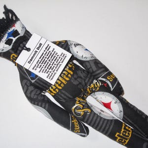 Puede incluir: Una mu&ntilde;eca de tela negra y amarilla con un logotipo de Steelers negro y blanco. La mu&ntilde;eca tiene una etiqueta que dice "Dammit Doll" y explica c&oacute;mo usar la mu&ntilde;eca para desahogar la frustraci&oacute;n.
