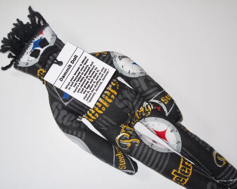 Puede incluir: Una mu&ntilde;eca de tela negra y amarilla con un logotipo de Steelers negro y blanco. La mu&ntilde;eca tiene una etiqueta que dice "Dammit Doll" y explica c&oacute;mo usar la mu&ntilde;eca para desahogar la frustraci&oacute;n.