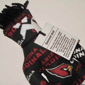 May include: A black and red Arizona Cardinals "Dammit Doll" with black fringe hair. The doll features the team's logo and name printed on the fabric. A tag attached to the doll provides instructions on how to use it.