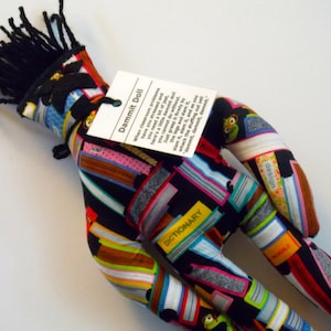 May include: A black and multi-colored fabric doll with a black yarn afro hair. The doll is made of fabric with a pattern of books and the words "Dammit Doll", "Dictionary", "World", and "design".