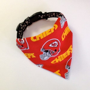 Puede incluir: Bandana roja y amarilla para perro con el logotipo de los Kansas City Chiefs y la palabra "Chiefs" repetida en la bandana. La bandana est&aacute; unida a un collar de perro negro con huellas de patas blancas.