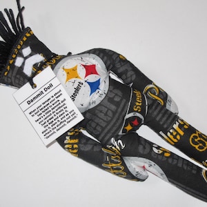 Puede incluir: Una mu&ntilde;eca "Dammit Doll" con tem&aacute;tica de los Pittsburgh Steelers, de color negro y amarillo, con una franja negra y blanca en la cabeza. La mu&ntilde;eca est&aacute; hecha de tela y tiene las palabras "Dammit Doll" y una descripci&oacute;n de c&oacute;mo usar la mu&ntilde;eca impresa en una etiqueta blanca unida a la mu&ntilde;eca.