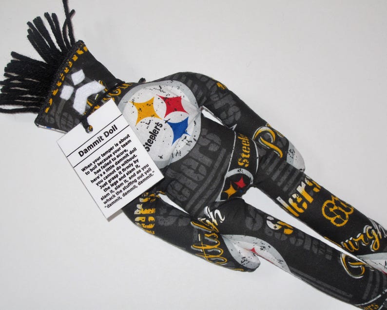 Puede incluir: Una mu&ntilde;eca "Dammit Doll" con tem&aacute;tica de los Pittsburgh Steelers, de color negro y amarillo, con una franja negra y blanca en la cabeza. La mu&ntilde;eca est&aacute; hecha de tela y tiene las palabras "Dammit Doll" y una descripci&oacute;n de c&oacute;mo usar la mu&ntilde;eca impresa en una etiqueta blanca unida a la mu&ntilde;eca.