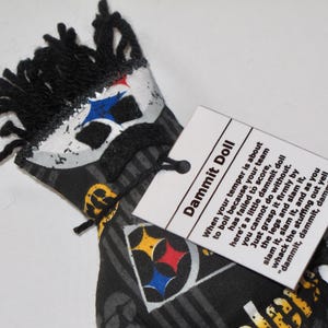 Puede incluir: Una mu&ntilde;eca de tela negra y blanca con un logotipo de los Pittsburgh Steelers y una etiqueta que dice "Dammit Doll" con instrucciones sobre c&oacute;mo usar la mu&ntilde;eca para desahogar la frustraci&oacute;n.
