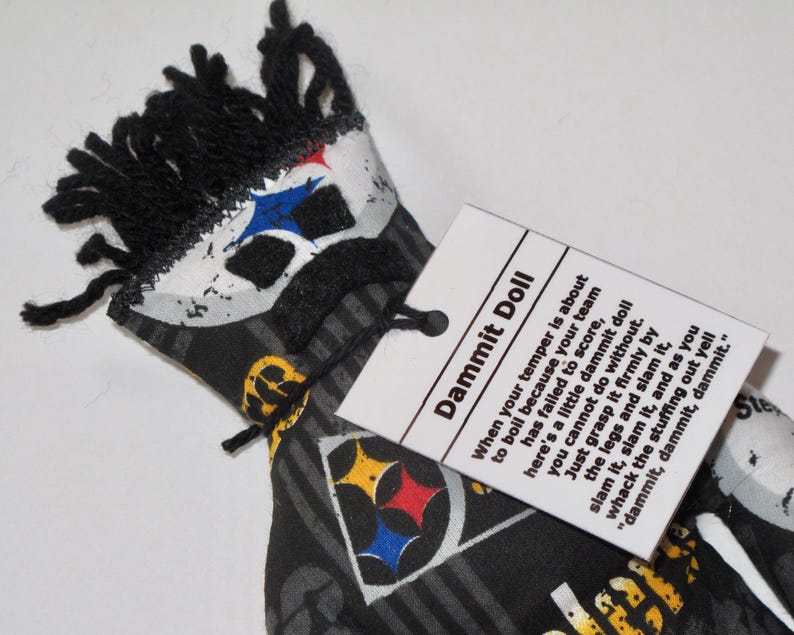 Puede incluir: Una mu&ntilde;eca de tela negra y blanca con un logotipo de los Pittsburgh Steelers y una etiqueta que dice "Dammit Doll" con instrucciones sobre c&oacute;mo usar la mu&ntilde;eca para desahogar la frustraci&oacute;n.