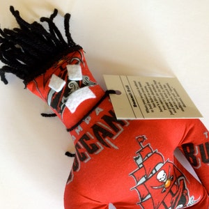 May include: Red and black fabric doll with a pirate ship design and the words "Tampa Bay Buccaneers" on the fabric. The doll has black yarn hair and white felt eyes. The doll is holding a tag that says "Dammit Doll" and has instructions on how to use the doll.