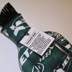May include: A handmade "Dammit Doll" in the shape of a hand, featuring a green and white pattern with the text "JETS". The doll has black fringe at the top and a tag with instructions. The tag reads "Dammit Doll".