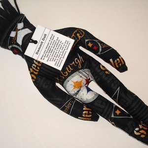 Puede incluir: Una mu&ntilde;eca de tela negra y amarilla con una franja negra en la cabeza. La mu&ntilde;eca lleva una camiseta de los Pittsburgh Steelers y tiene una etiqueta que dice "Dammit Doll" con instrucciones sobre c&oacute;mo usar la mu&ntilde;eca para desahogar la frustraci&oacute;n.