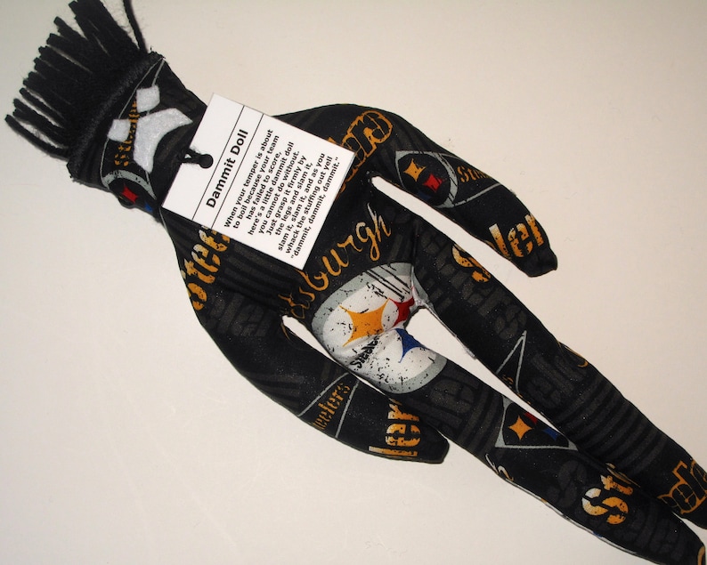 Puede incluir: Una mu&ntilde;eca de tela negra y amarilla con una franja negra en la cabeza. La mu&ntilde;eca lleva una camiseta de los Pittsburgh Steelers y tiene una etiqueta que dice "Dammit Doll" con instrucciones sobre c&oacute;mo usar la mu&ntilde;eca para desahogar la frustraci&oacute;n.