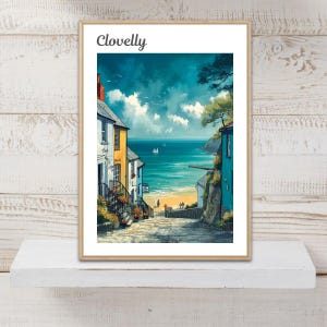 May include: A framed art print titled Clovelly featuring a coastal village scene with colourful houses, a stone path leading to a sandy beach, and a blue sea under a cloudy sky. The print displays a vibrant landscape with sailboats on the water.