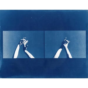 May include: A cyanotype print showing two panels, each with a pair of hands in a gesture of connection. The hands are white against a blue backdrop, creating a striking contrast. The image is framed by a dark blue border.