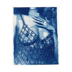 Może przedstawiać: Niebieski nadruk topu z kolczugi. Top wykonany jest z małych, połączonych ze sobą metalowych ogniw, tworzących odzież przypominającą biustonosz. Dłoń z długimi paznokciami spoczywa na piersi. Obraz ma estetykę vintage.