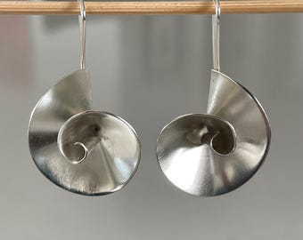 May include: A pair of silver-colored earrings with a spiral design. Each earring has a hook for pierced ears and a half-moon shape that curves into a spiral. The earrings are hanging from a wooden dowel against a neutral background.
