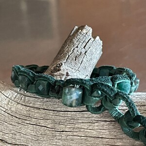 May include: A green leather bracelet with dark green gemstone beads. The bracelet is wrapped around a piece of driftwood.