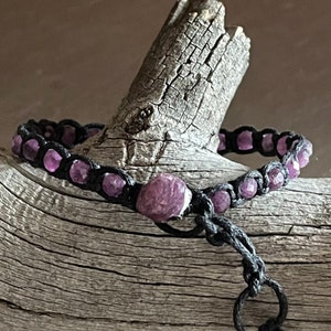 May include: A purple gemstone bracelet with black cord and a sliding knot closure. The bracelet is made with small, faceted purple gemstones and a larger, round purple gemstone.