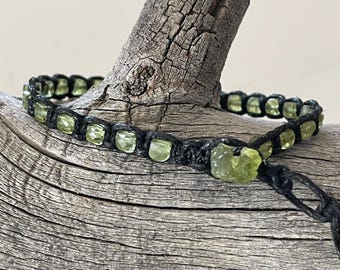 Puede incluir: Un brazalete de piedras preciosas verdes con cordón negro. El brazalete está hecho con pequeñas piedras preciosas de forma cuadrada y está atado con un nudo en el extremo.