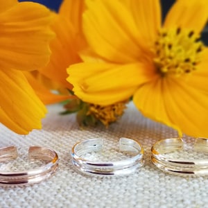 May include: Three silver toe rings with a double band design. The rings are on a light brown fabric background.