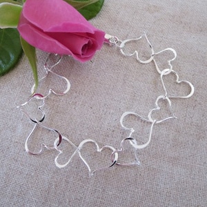 May include: A silver bracelet made of linked heart shapes. The bracelet is on a light brown fabric background.