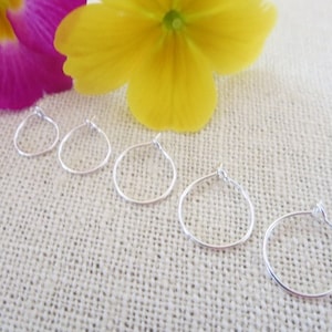 May include: Five silver hoop earrings of varying sizes, arranged in a row on a white textured surface.