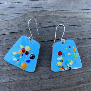 May include: A pair of blue enamel earrings with a silver hook. The earrings are shaped like a trapezoid and have a colorful confetti pattern.