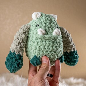 May include: A green, gray, and teal crocheted stuffed animal with a white nose and ears. The animal is being held by a hand.