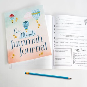 Op de afbeelding: Een roze en blauw dagboek met een aquarel-illustratie van luchtballonnen en sterren. De omslag heeft de tekst "Five Minute Jummah Journal". De binnenpagina's bevatten prompts voor reflectie en dankbaarheid na het vrijdaggebed.