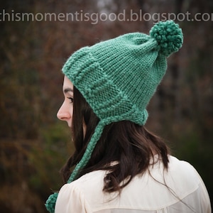 Könnte beinhalten: Eine grüne Strickmütze mit einem Bommel oben und Ohrenklappen. Die Mütze wird von einer Person mit langen braunen Haaren getragen.