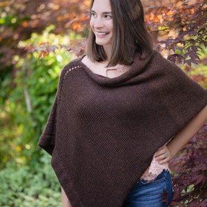 May include: A brown knitted poncho with a cowl neck. The poncho is worn by a person with long brown hair and is standing in front of a leafy background.