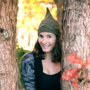 Op de afbeelding: Een vrouw met lang bruin haar draagt een groene gebreide muts met een puntige top. Ze glimlacht en kijkt naar de camera. De muts is gemaakt van een getextureerd garen en heeft een unieke vorm.