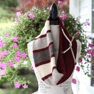 May include: A knitted infinity scarf displayed on a mannequin. The scarf features a striped pattern in cream, gray, and burgundy. The other side is solid burgundy with button closures and cream-colored ties. The background includes pink flowers.